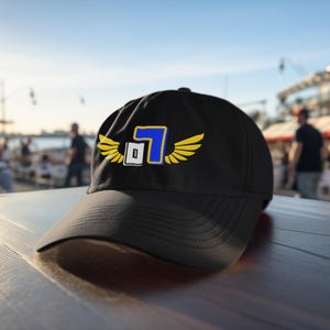 Puede incluir: Gorra de béisbol negra con un diseño bordado en amarillo y azul que presenta un número "7" con alas. El número "7" está dentro de un cuadrado blanco.
