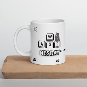 Puede incluir: Taza de cerámica blanca con un gato negro sosteniendo una taza de café. La taza tiene texto negro que dice "W A S D NESDAY" y un diseño de grano de café.
