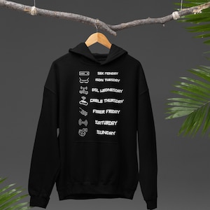 Op de afbeelding: Zwarte hoodie met witte graphics en tekst. De tekst bevat de dagen van de week met bijbehorende internetgerelateerde pictogrammen. De vermelde dagen zijn maandag tot en met zondag. De hoodie hangt aan een houten hanger.