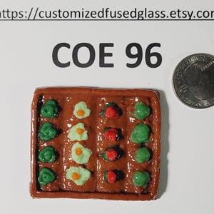 May include: A miniature fused glass garden scene with green, red, and yellow details. The brown rectangular piece features rows of colorful glass vegetables and eggs. The text "COE 96" is above the garden. A quarter is next to the garden.