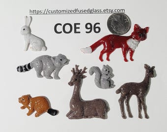 May include: A collection of seven small animal-shaped fused glass pieces including a white rabbit, a red fox, a gray raccoon, a brown beaver, a gray squirrel, a brown deer, and a brown fawn, with a US quarter and the text COE 96 for scale.