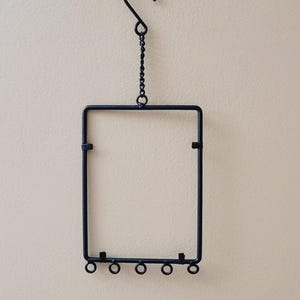May include: Black metal hanging frame with a rectangular shape, suspended from a hook and chain. The frame has small circular details at the bottom. The background is a neutral beige color.
