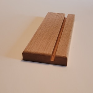 May include: A simple wooden phone stand with a slot for holding a phone upright. The stand is made of light brown wood and has a smooth finish.