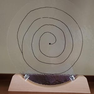 May include: A clear glass disc with a black spiral drawn on it, mounted on a wooden base. The base has two silver metal screws on each side.