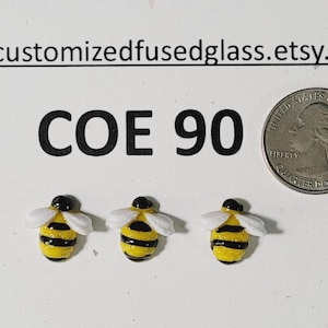 May include: Three small, handmade glass bee figurines with yellow and black striped bodies, white wings, and black heads. A US quarter coin is next to the bees for size comparison. The text "COE 90" is above the bees.