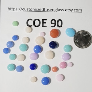 May include: A collection of colorful glass beads in various sizes and shades, including blue, green, pink, and orange. The text "COE 90" is displayed, along with a website address. A U.S. quarter coin is included for size reference.
