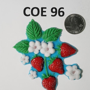 May include: A fused glass strawberry design featuring red strawberries, white flowers, and green leaves on a blue base. The text "COE 96" is above the design. A coin is next to the design for size reference.