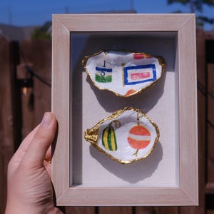 May include: Two oyster shells painted with nautical designs are displayed in a gold frame. One shell features a red, white, and blue flag with the number "156" on it. The other shell has a green and yellow gourd and a red and white buoy.