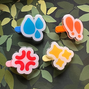 May include: Four colorful hair clips with different designs. One clip features blue water droplets, another has orange bunny ears, a third shows a red angry face, and the last has yellow stars. Each clip has a different colored base.
