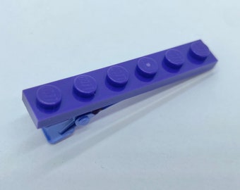 May include: A purple plastic hair clip shaped like a Lego brick with five studs. The clip is made to look like a Lego brick.