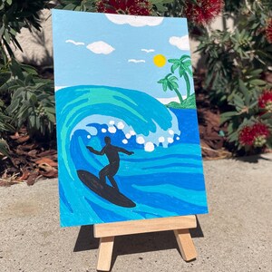 Op de afbeelding: Een schilderij op canvas dat een surfer toont die op een blauwe golf met wit schuim rijdt. De achtergrond toont een blauwe lucht met witte wolken, een gele zon en palmbomen op een klein eiland.
