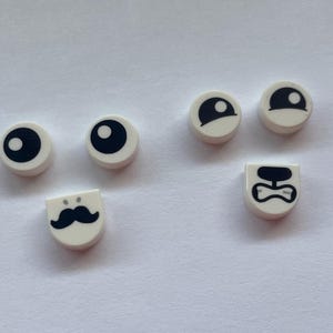 May include: A collection of white, circular and rectangular Lego pieces with black graphic designs. The pieces feature eyes, a mustache, and a bow tie, suggesting a playful or character-building theme.