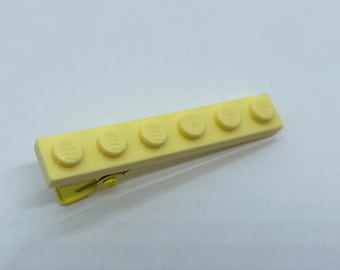 May include: A yellow plastic hair clip shaped like a Lego brick with six studs. The clip is made of a single piece of plastic and has a gold metal spring clip.