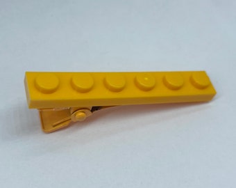 May include: A yellow plastic hair clip shaped like a Lego brick with six studs. The clip is made of a single piece of plastic and has a metal spring clip on the back.