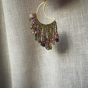 Pode incluir: Um brinco de lua crescente dourado com franjas de contas em tons de verde, roxo e transparente. O brinco tem um estilo boêmio delicado, com pequenas contas criando um efeito pendurado e texturizado.