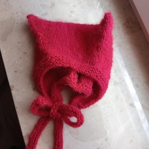Könnte beinhalten: Ein leuchtend roter, gestrickter Babyhut mit spitzen Ohren und einem Schleifenverschluss. Der Hut ist aus weichem, strukturiertem Garn gefertigt. Dieser Hut ist entworfen, um den Kopf eines Babys warm zu halten. Die Farbe ist ein leuchtendes Rot.