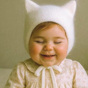 Peut inclure: Un bébé souriant portant un bonnet blanc en tricot avec des oreilles de chat et une robe à imprimé floral avec un col noué. Les yeux du bébé sont fermés et les joues sont roses.