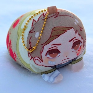 Puede incluir: Un juguete de peluche cilíndrico con una cara de dibujos animados, cabello castaño y una cadena dorada. El juguete está decorado con patrones florales rojos y blancos y está sobre una superficie nevada.