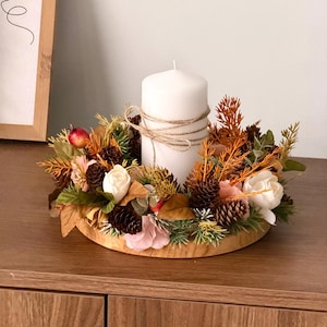 May include: A white pillar candle wrapped in twine sits in the centre of a circular wooden base adorned with a decorative wreath of pinecones, dried orange foliage, green sprigs, and pale pink and white artificial flowers.