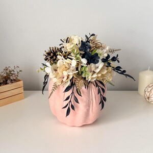 Puede incluir: Un recipiente con forma de calabaza de color rosa claro lleno de un arreglo de flores color crema, follaje azul marino oscuro y piñas marrones. El arreglo floral se asienta sobre la calabaza, con algunas hojas oscuras colgando por los lados.