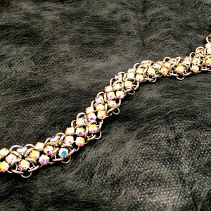 Może przedstawiać: Srebrna bransoletka z łańcuszkiem ozdobiona tęczowymi kryształami. Bransoletka jest wykonana w splocie kolczugi i ma zapięcie.