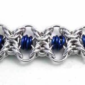 May include: Close-up of a silver chainmail bracelet with blue coiled accents. The bracelet is made of interlocking metal rings and features a repeating pattern of silver and blue elements. The design is intricate and detailed.
