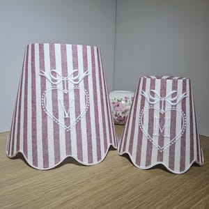 May include: Two striped lampshades in varying sizes. The shades feature a white and burgundy vertical stripe pattern. Each shade has a white embroidered heart with a letter and bow detail. The edges of the shades have a scalloped design.
