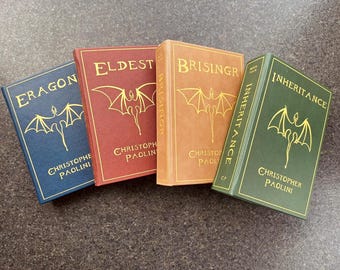 May include: Four hardcover books with dragon illustrations and gold lettering. The titles are "Eragon", "Eldest", "Brisingr", and "Inheritance" by Christopher Paolini.