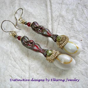 May include: A pair of dangle earrings featuring a dark, textured, elongated clay component with red accents, a small red bead, and a gold-toned wire-wrapped green stone. The base consists of a white, teardrop-shaped shell with a yellow band.