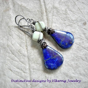 May include: A pair of drop earrings featuring teardrop-shaped blue lapis lazuli stones with gold flecks, accented by small square pale green beads. The stones are wrapped in dark oxidized silver wire with copper details and hanging hooks.