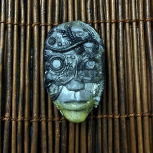 May include: A sculpted mask with a cyborg design, featuring metallic gray and black details. The mask has a human-like face with a green-tinged lower portion. The background is a woven bamboo mat.
