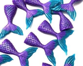 May include: A collection of purple and teal mermaid tail decorations. The tails are detailed with a scale pattern and glitter accents. The mermaid tails are arranged on a white background.