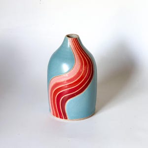 May include: A light blue ceramic vase with a wavy, abstract design in shades of pink and red.