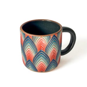 May include: A ceramic mug with a black handle and a colorful, geometric pattern. The pattern features blue, red, and pink shapes. The interior of the mug is dark blue, and the rim is brown.