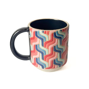 May include: A ceramic mug with a black handle and interior. The exterior features a repeating wavy pattern in shades of red, pink, blue, and light blue. The mug sits on a white surface.