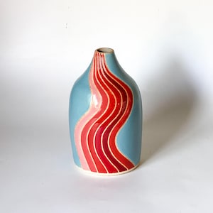 May include: A light blue ceramic vase with a wavy, abstract design in shades of pink and red.
