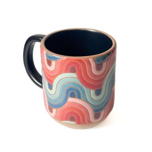 May include: A ceramic mug with a black handle and a colorful, wavy pattern in shades of red, pink, and blue. The interior is dark blue. The mug is a unique piece of drinkware.