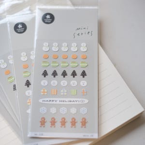 May include: Several packs of clear stickers featuring winter-themed designs. The stickers include snowmen, mugs, Christmas trees, presents, snowflakes, and gingerbread men. The text "HAPPY HOLIDAY!" is printed on one of the stickers. The stickers are labeled "mini series".