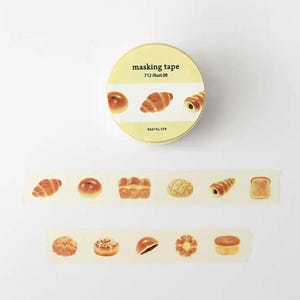May include: A yellow masking tape roll featuring illustrations of baked goods like croissants and bread rolls. The tape is labelled "masking tape 712 illust.08". Unrolled tape shows more bread-themed illustrations, perfect for craft projects.