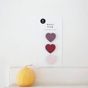 May include: A white card with the text "DAILY PLAN" and three heart-shaped sticky notes in shades of purple, red, and pink. A yellow, lemon-shaped candle sits below the card.