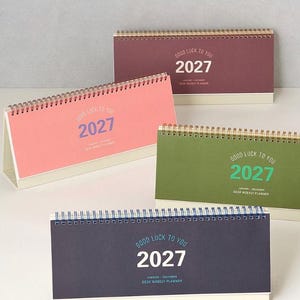 May include: Four horizontal desk weekly planners for the year 2027, each featuring a spiral binding at the top. The planners come in maroon, pink, olive green, and dark blue, with the text "GOOD LUCK TO YOU 2027" and "JANUARY-DECEMBER DESK WEEKLY PLANNER".