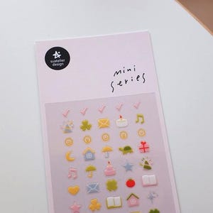 Puede incluir: Una hoja de pegatinas con varios diseños en miniatura coloridos. Las pegatinas incluyen una marca de verificación, un trébol de cuatro hojas, un sobre, una vela, una nota musical, una moneda, una luna creciente, una casa, un paraguas, una tortuga, una cruz y más. El texto "mini series" está escrito en la hoja.