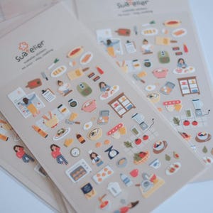 May include: Sheets of decorative stickers featuring cooking-themed illustrations. The stickers include images of kitchen tools, food items, and people cooking. The color palette includes soft pastels and earth tones, with a focus on a cooking theme.