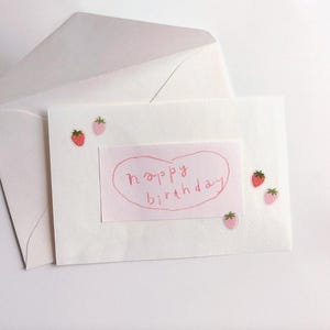 May include: A white greeting card with a pink heart-shaped sticker that says "happy birthday" in handwritten script. The card is decorated with small strawberry stickers and is placed in a white envelope.