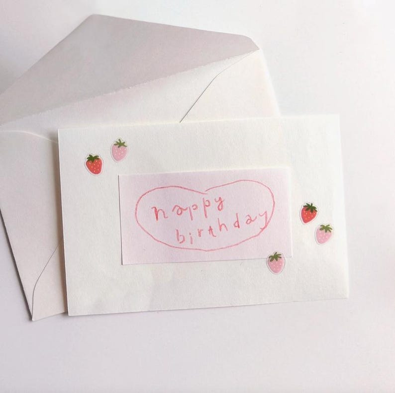 May include: A white greeting card with a pink heart-shaped sticker that says "happy birthday" in handwritten script. The card is decorated with small strawberry stickers and is placed in a white envelope.