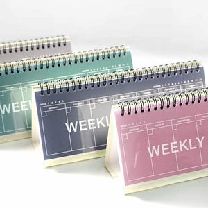 Puede incluir: Calendarios de escritorio con encuadernación en espiral. Presentan un diseño de planificador semanal con la palabra "WEEKLY" en blanco. La paleta de colores incluye tonos azules, verdes y rosas. Ideales para la planificación semanal.