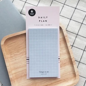 Puede incluir: Un bloc de notas adhesivas azul claro con un patrón de cuadrícula, etiquetado como "Things to do" y "Daily Plan". El bloc de notas está sobre un fondo rosa con el logotipo "Suatelier Design". El bloc de notas está sobre una bandeja de madera.
