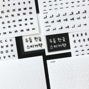 May include: A collection of white sticker sheets with black Korean Hangul characters and symbols. Some sheets have individual characters, while others have words. The text "두들 한글 스티커팩" is visible on a black and white label.