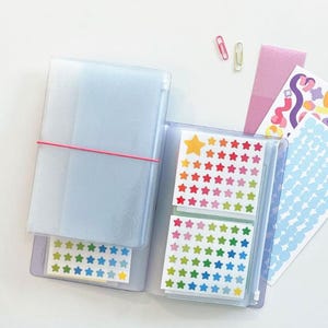 May include: Clear plastic sticker storage binder with a red elastic band, holding sheets of colourful star-shaped stickers. Additional stickers and paperclips are scattered nearby. The binder is designed for organising and storing stickers.
