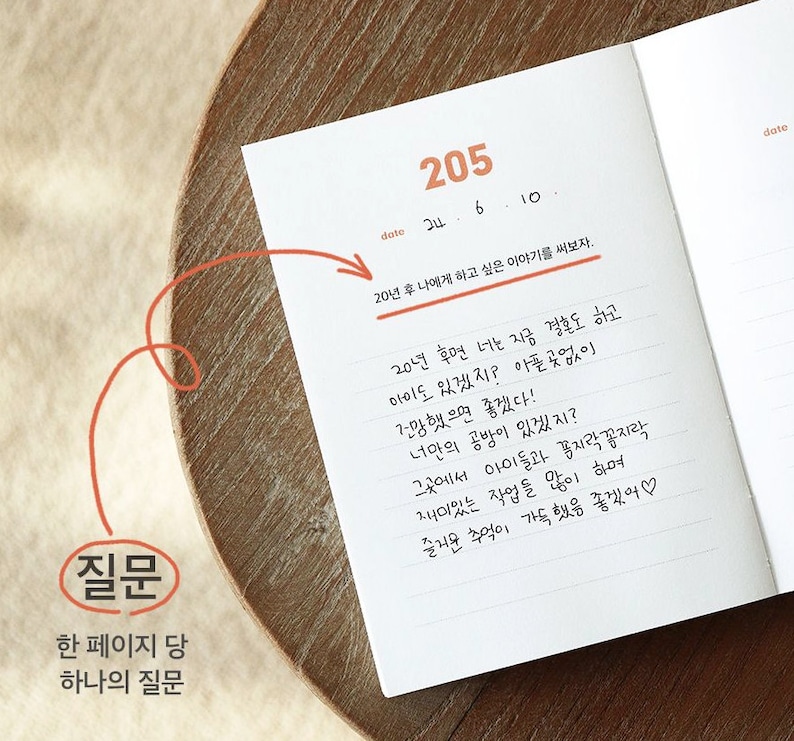 May include: A white notebook with a handwritten question prompt: "20 years from now, what would you tell your younger self?" The notebook is open to a page with the number 205 written at the top. The date is written as 24.6.10. The notebook is on a wooden surface.